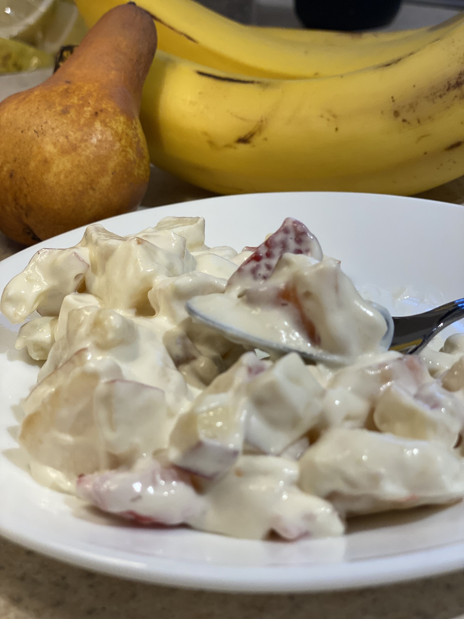 fruit salad with whipped topping on a white plate with a silver spoon and bananas and pears in the background.