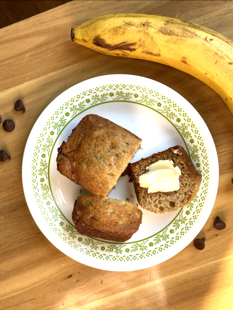 small loaf of banana bread, sliced and topped with butter, sitting on a small plate with green decorations around the outside, sitting on a table with chocolate chips and a banana