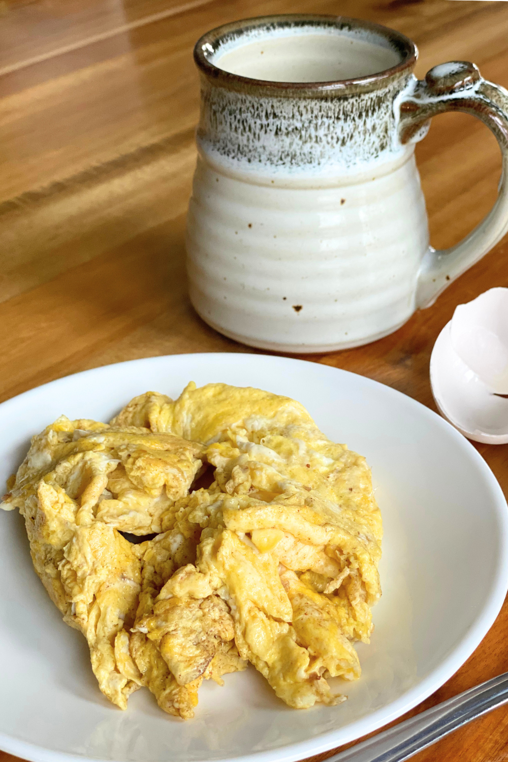 scrambled eggs on a white plate on a table next to a white mug and a cracked egg shell