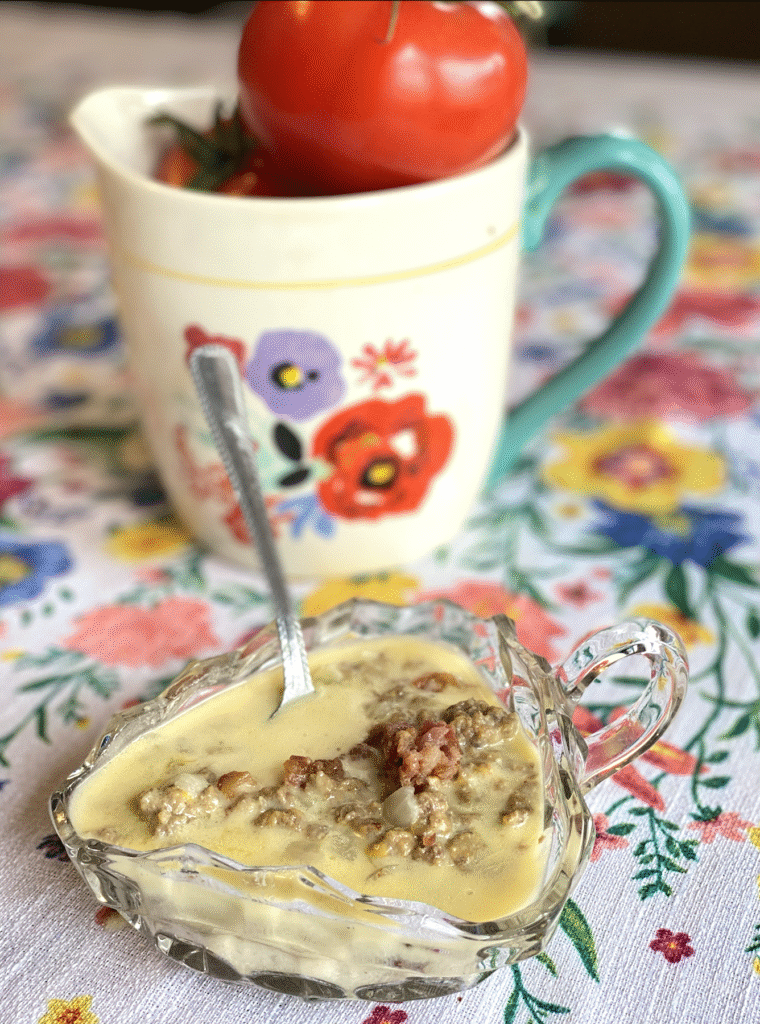 glass bowl of cheeseburger soup with a metal spoon on a floral tablecloth. A white pitcher with flower on it and a tomato inside in the background